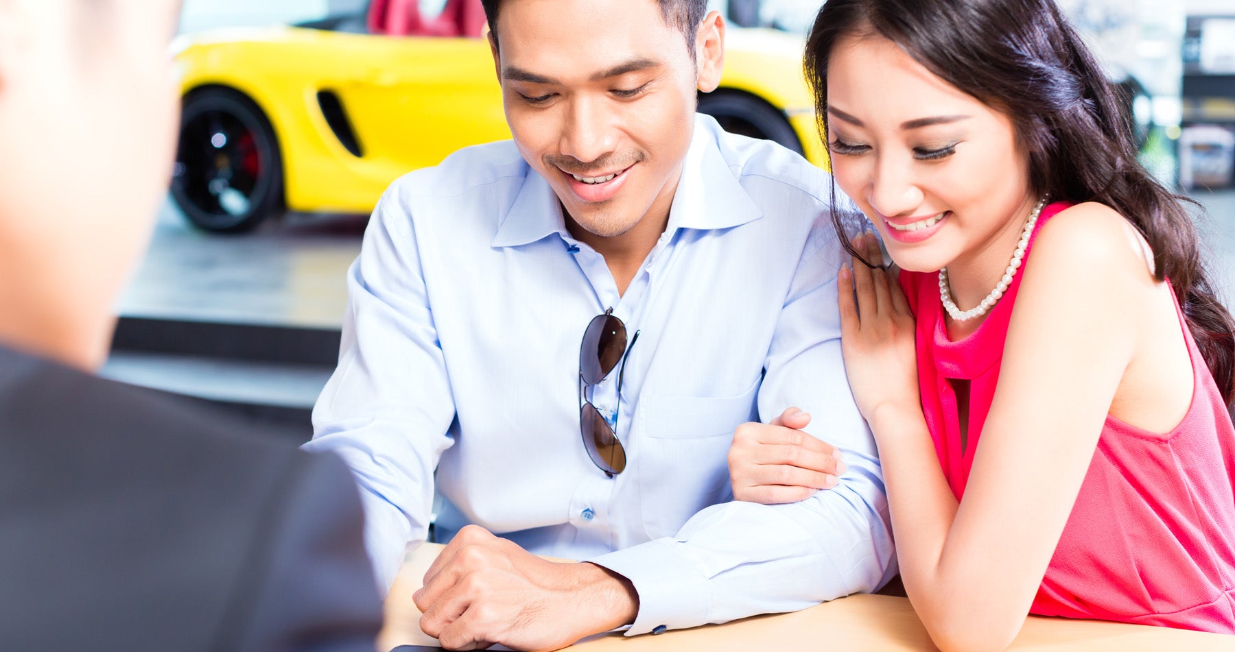A smiling couple, a woman in a pink dress and a man in a blue shirt, listening to a salesperson.