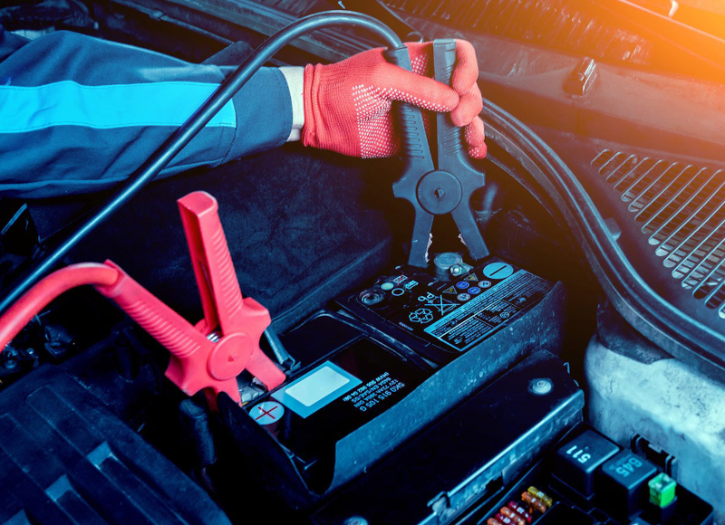 A person wearing red gloves connects black jumper cables to a car battery while a red cable is already attached.